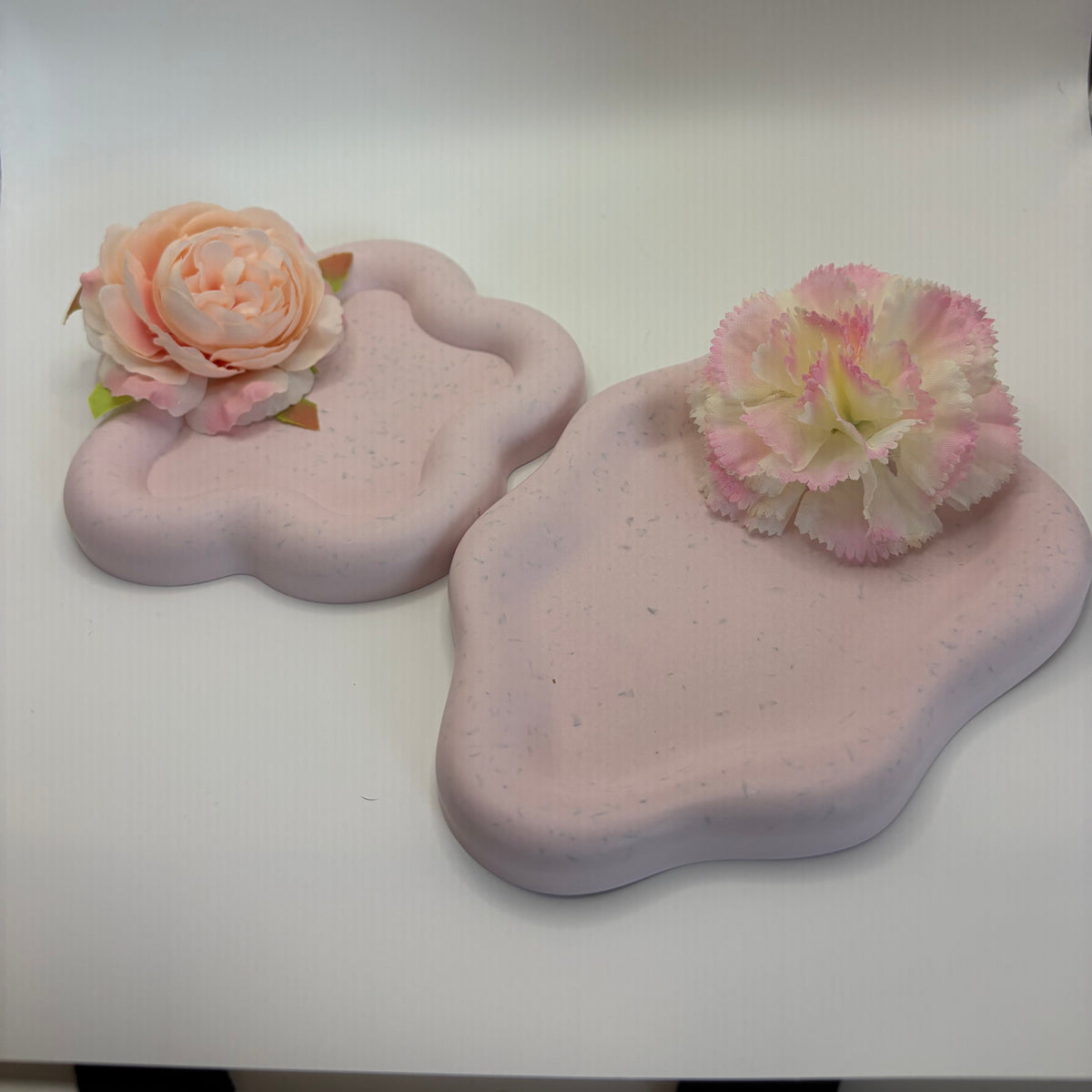 Two pastel pink cloud-shaped decorative trays or coasters with subtle speckles, each adorned with a fabric flower, displayed on a white studio background.