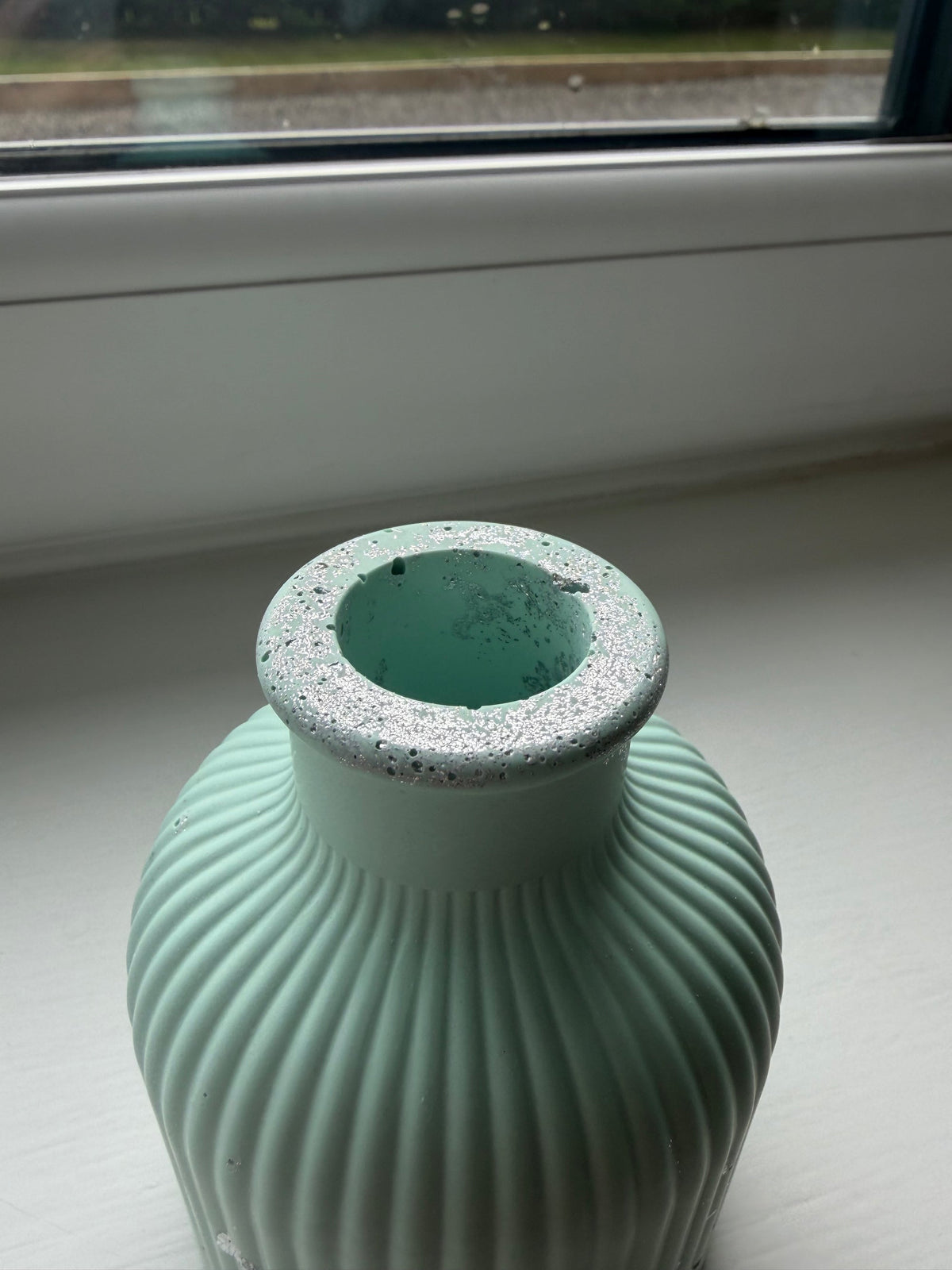 A ribbed jesmonite vase in a pale light green with silver leaf detailing and a speckled rim, photographed on a windowsill with a window and outdoor view in the background.