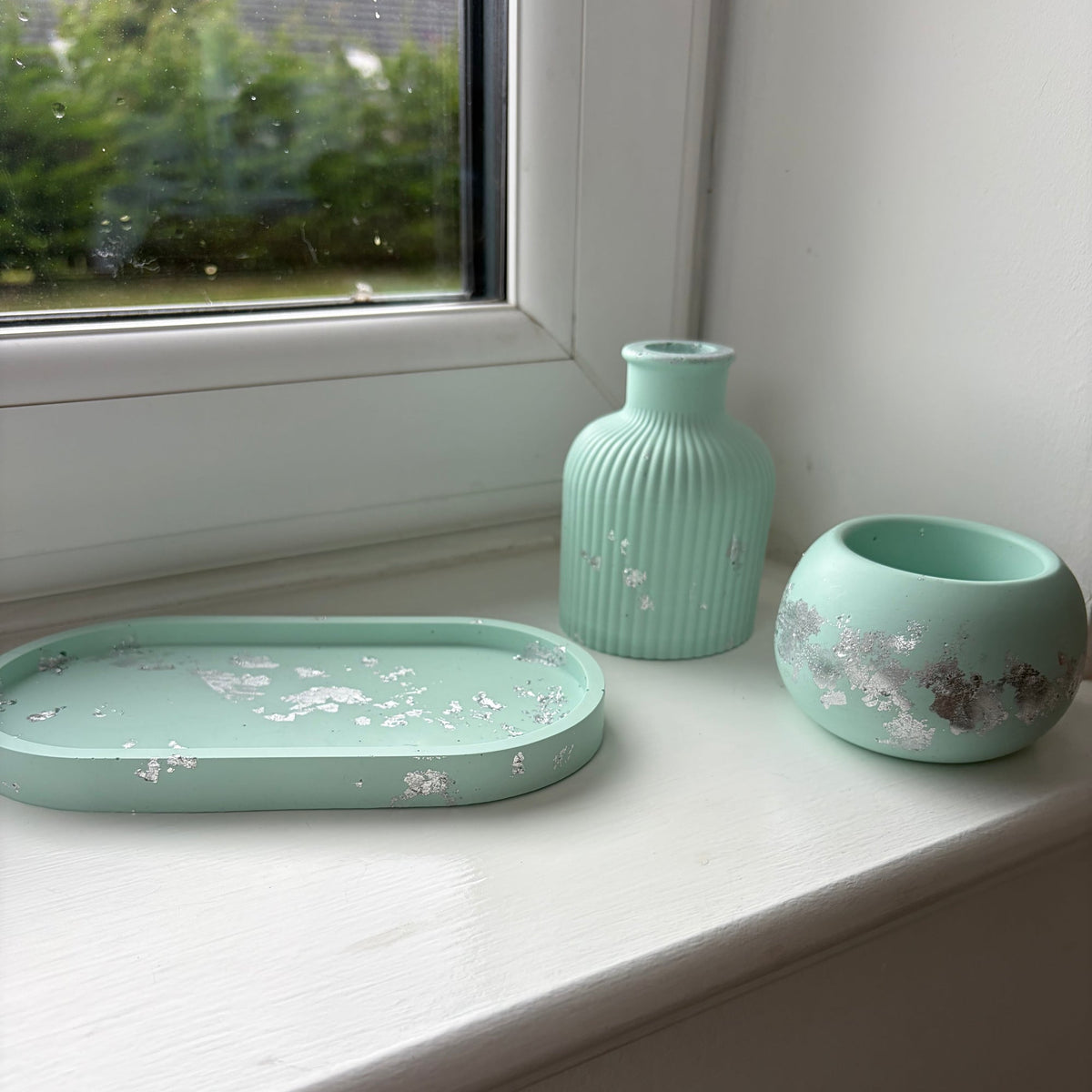 A set of three light green jesmonite home decor pieces with silver leaf accents, including a small ribbed vase, a round tealight holder, and an oval tray, arranged on a white windowsill.