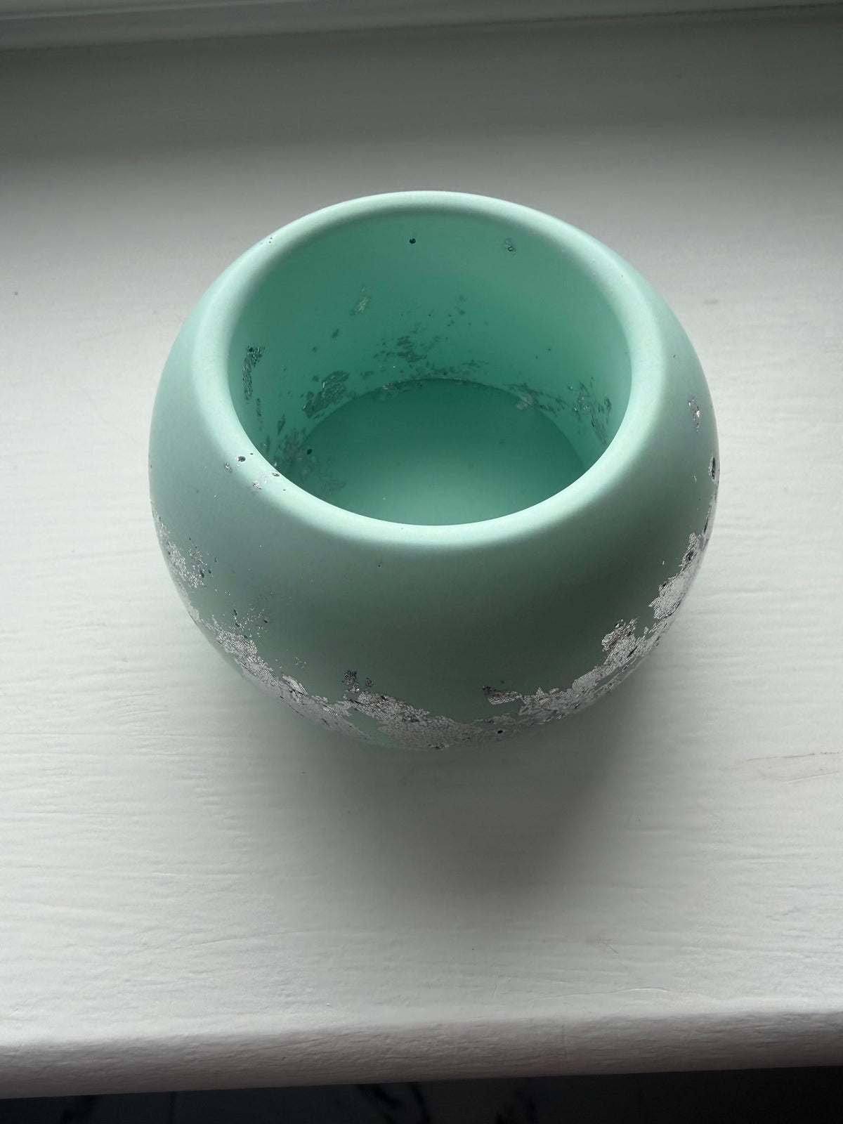 A small round jesmonite tealight holder in light green with a distressed silver leaf finish around the base, resting on a white wooden surface.