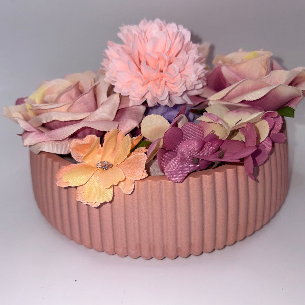 A pink ribbed ceramic planter filled with artificial pastel flowers, including a large pink chrysanthemum, peach roses, and purple hydrangeas, set against a plain light gray background.