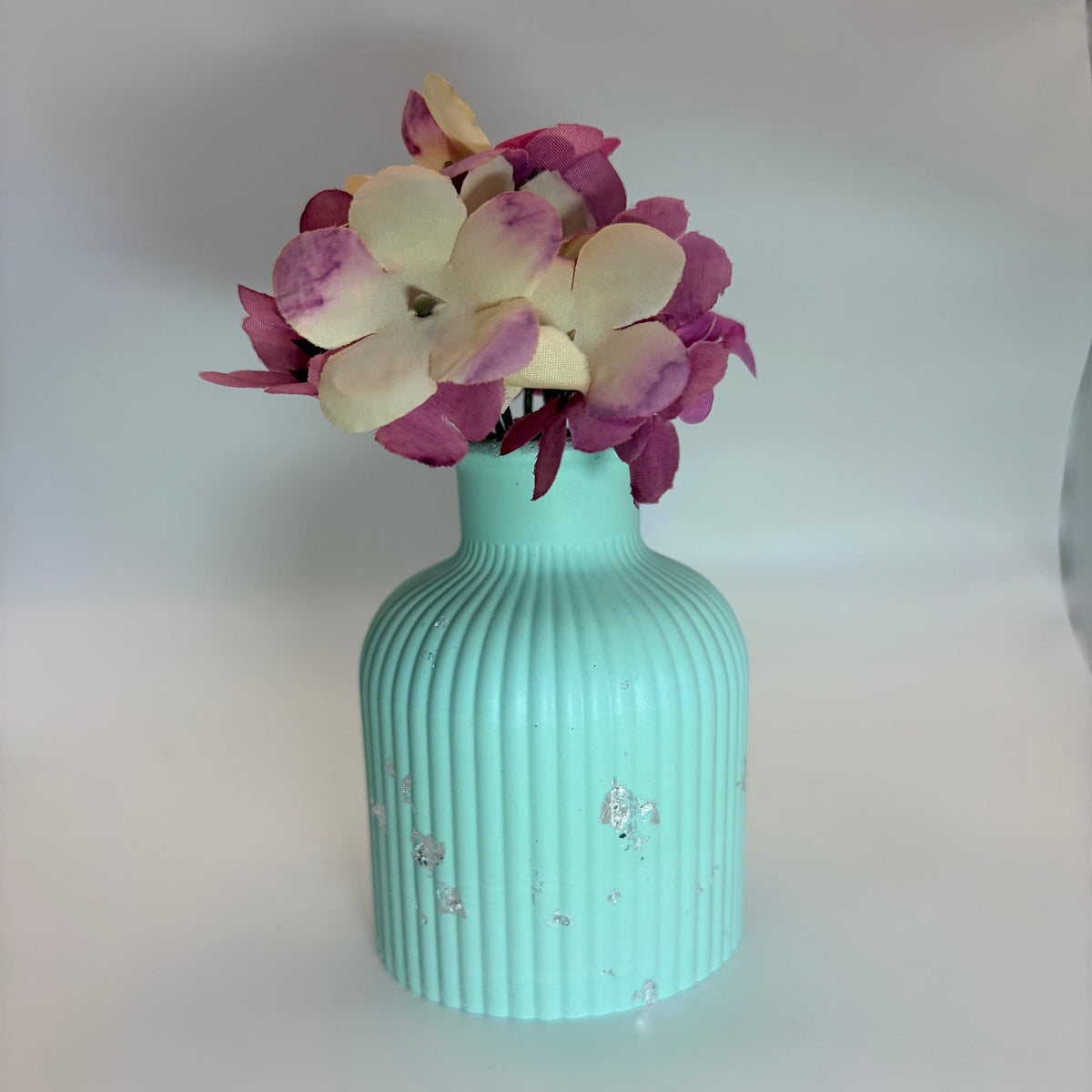 A mint green ribbed ceramic vase with small silver leaf accents, holding a bouquet of purple and cream hydrangea flowers, set against a plain light gray background.