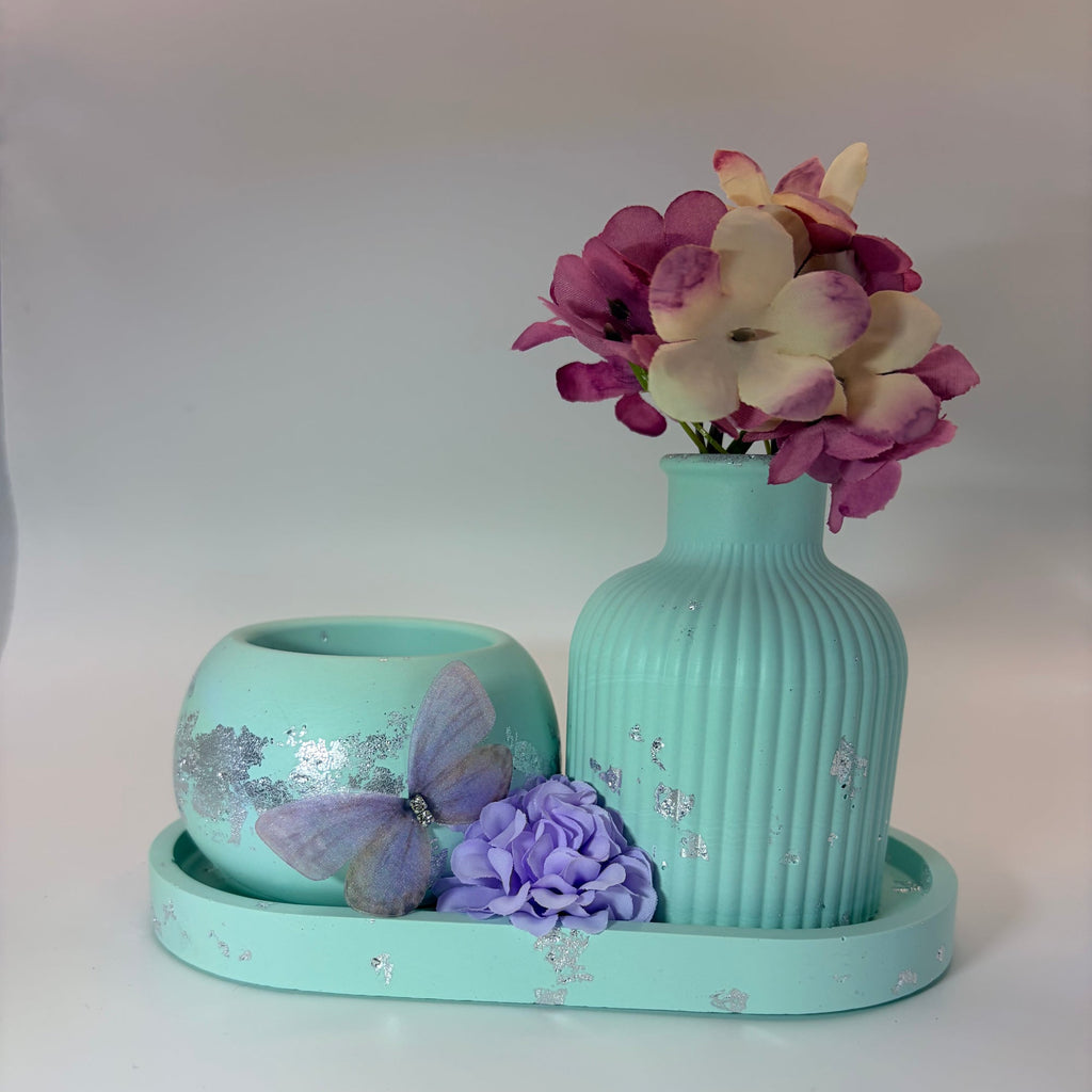 A decorative mint green ceramic set on a matching tray, featuring a ribbed vase with artificial pink and cream flowers, a small round bowl with a purple butterfly decoration, and a purple flower accent, all set against a plain light gray background.