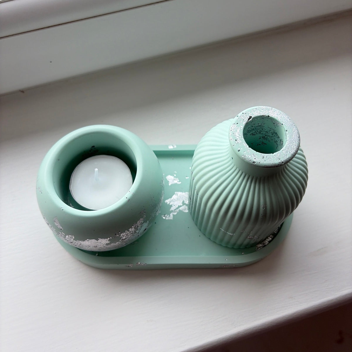 A light green jesmonite tealight holder and ribbed vase set on a white windowsill, featuring a round holder with a white tealight.