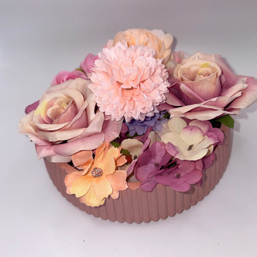 A round, ribbed pink planter box filled with an assortment of artificial flowers in pastel shades of pink, peach, purple, and cream, set against a plain light gray background.
