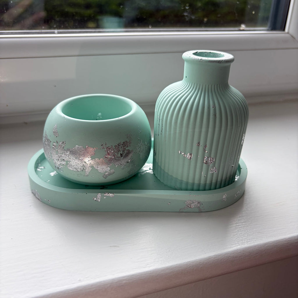 A jesmonite set in light green with silver leaf detailing, comprising a ribbed vase, a small round tealight holder, and a matching tray, displayed on a white windowsill with a window view in the background.
