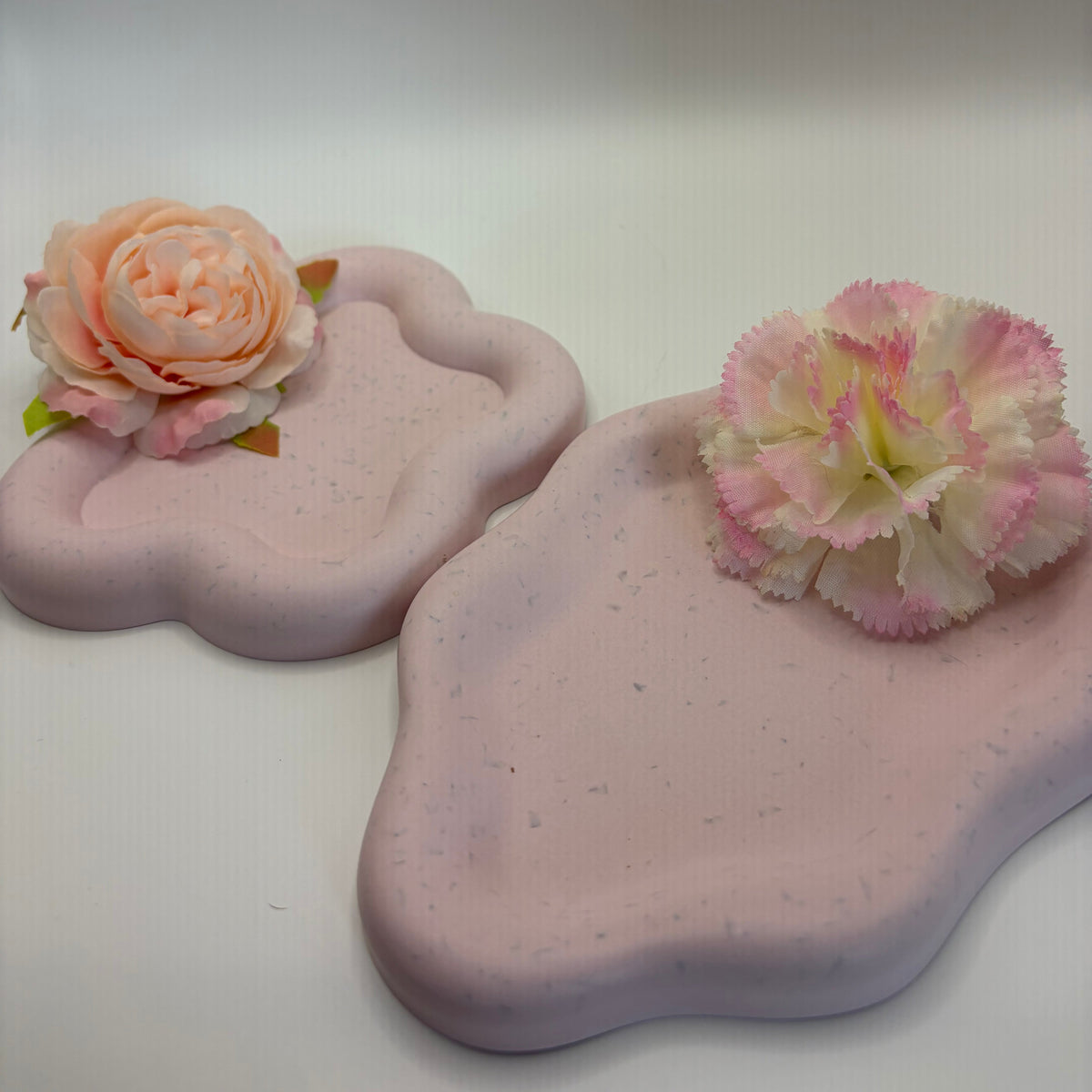 Two pastel pink cloud-shaped trays with subtle speckles, each decorated with a faux flower, set against a plain light gray background.