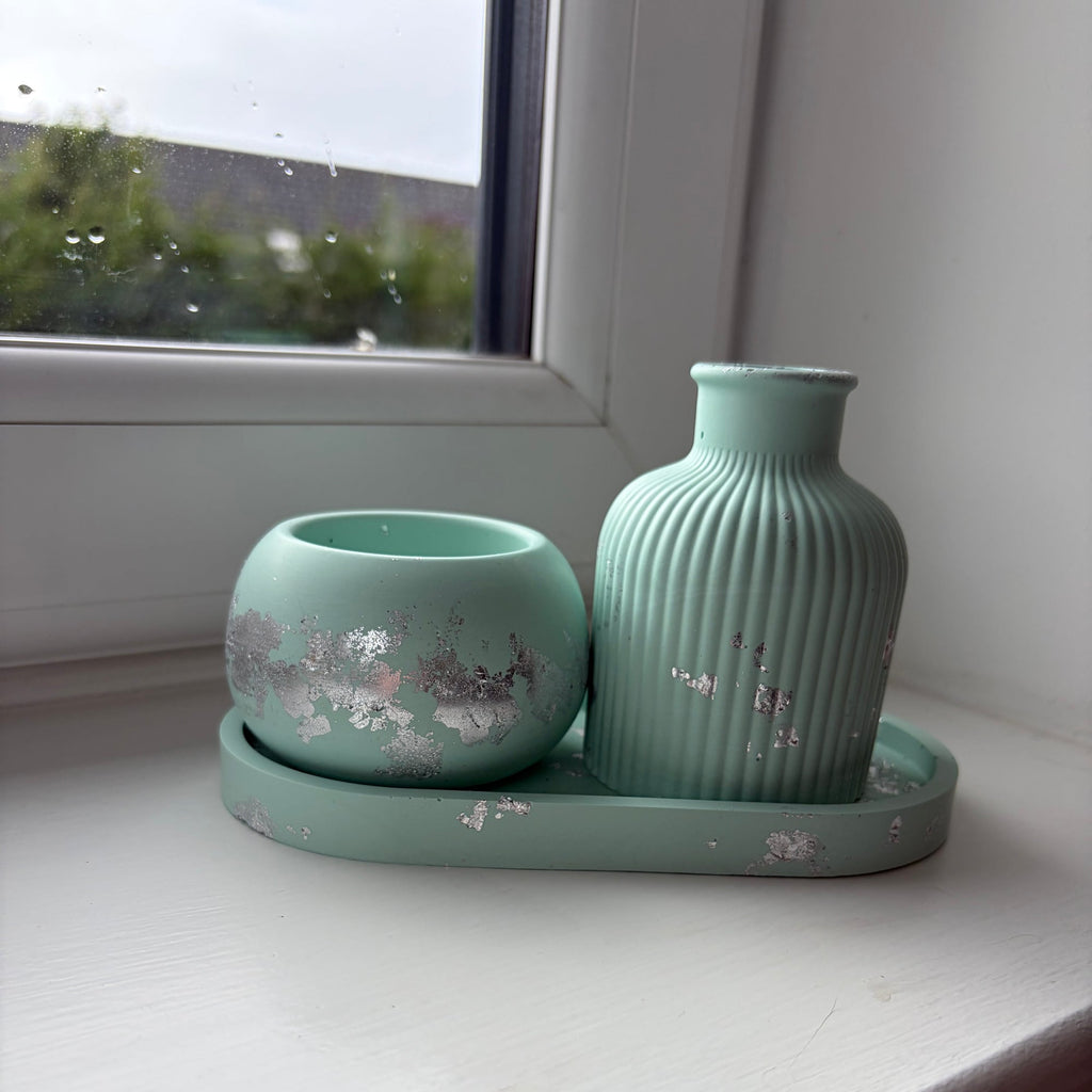 A light green jesmonite set consisting of a ribbed vase, a small tealight holder, and a matching tray, decorated with silver leaf flakes, displayed on a white windowsill.