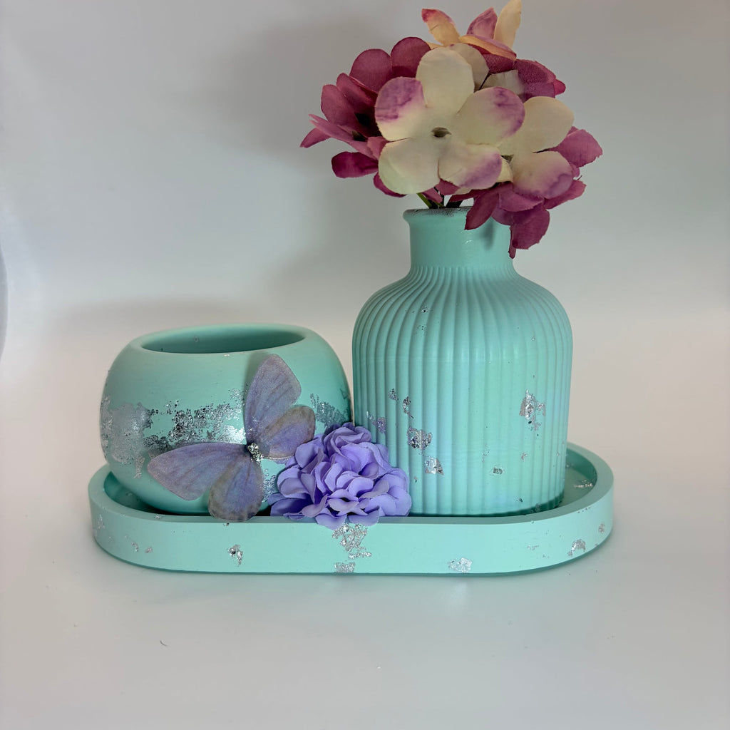 A mint green ceramic set on a matching tray, featuring a ribbed vase with pink and cream flowers, a small matching bowl with a purple butterfly decoration, and a purple flower accent, all on a white background.