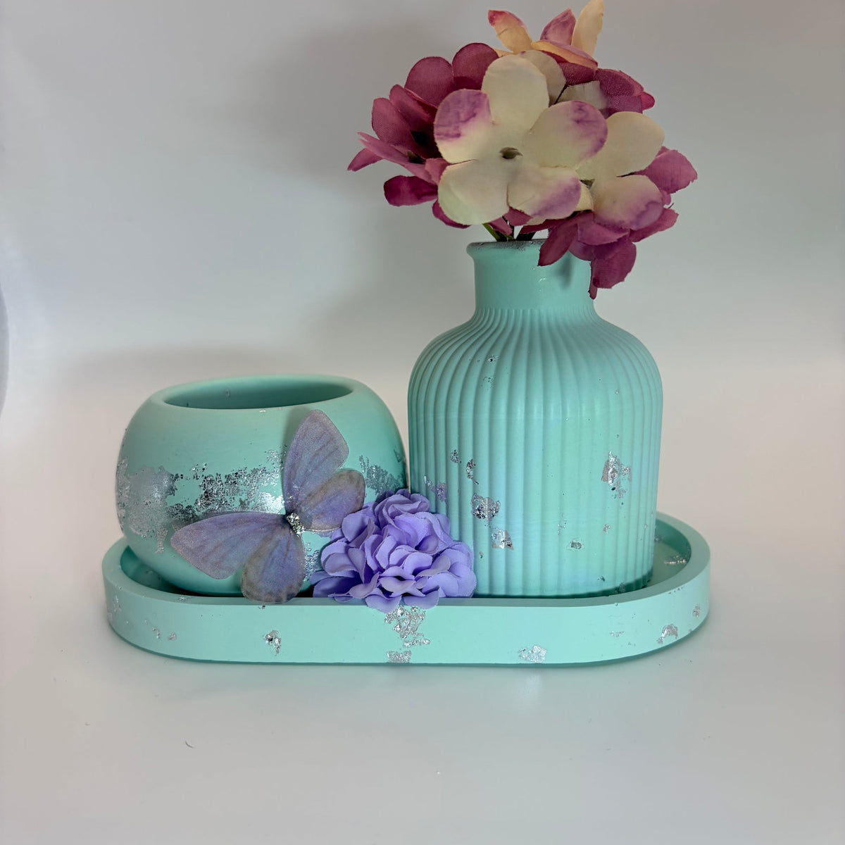 A mint green ceramic set on a matching tray, featuring a ribbed vase with pink and cream flowers, a small matching bowl with a purple butterfly decoration, and a purple flower accent, all on a white background.