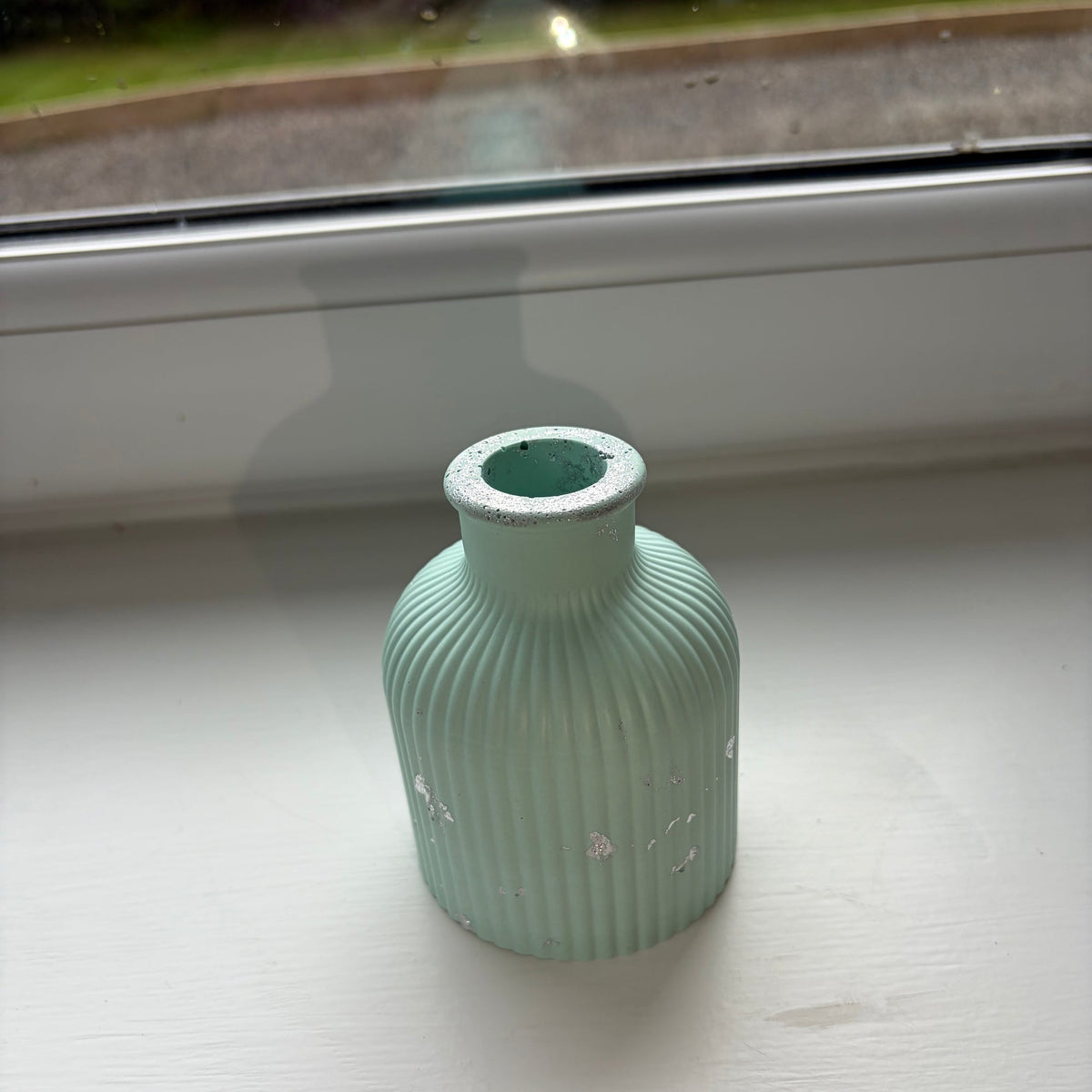 A small ribbed jesmonite vase in a pale light green with silver leaf accents, sitting on a white windowsill with a window view in the background.