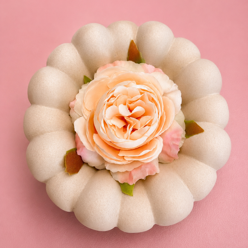 A decorative faux pumpkin with a scalloped cream exterior and a peach-pink rose flower in the center, set against a solid pink background.