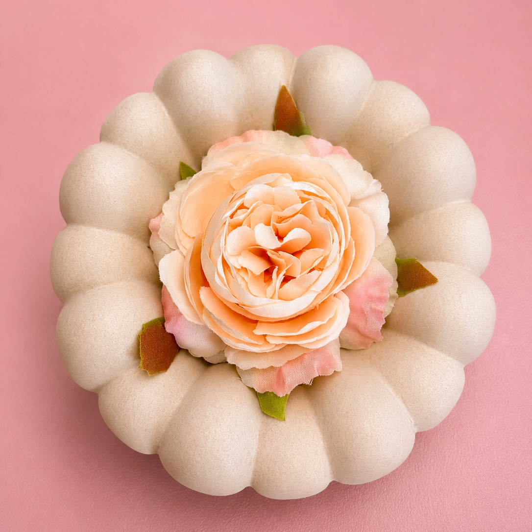 A decorative faux pumpkin with a scalloped cream exterior and a peach-pink rose flower in the center, set against a solid pink background.