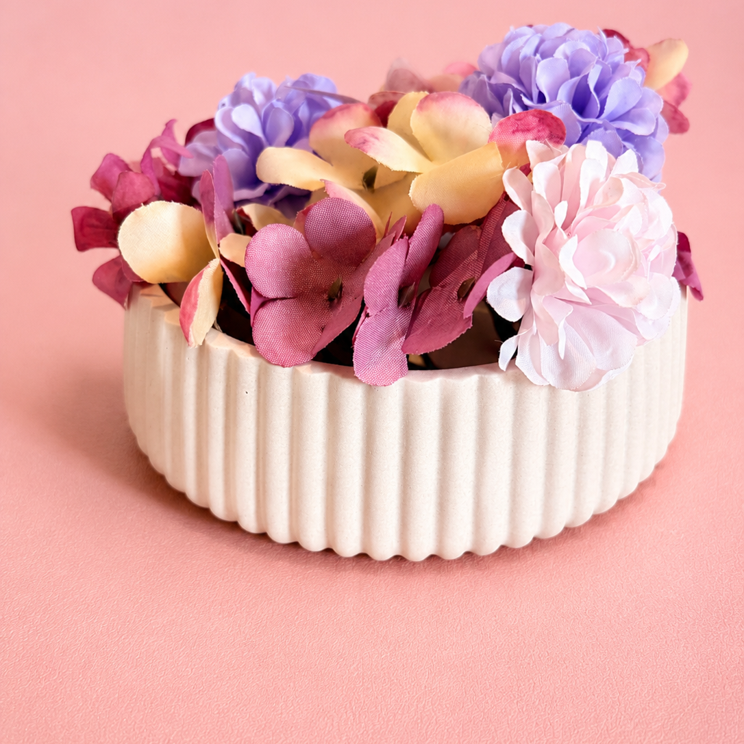 A collection of colorful artificial flowers in shades of purple, pink, and yellow, arranged in a white ribbed ceramic pot against a solid pink background.