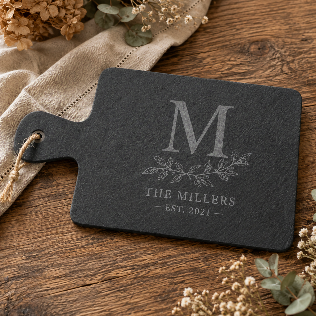 A personalized dark gray slate cutting board with a handle, engraved with a monogram 'M', floral motif, and 'THE MILLERS EST. 2021'.