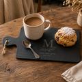 A black slate serving board on a rustic wooden table, styled with a ceramic mug of coffee, a spoon, and a scone dusted with powdered sugar. The board is engraved with a large monogram 'M', the name 'THE MILLERS', and 'EST.