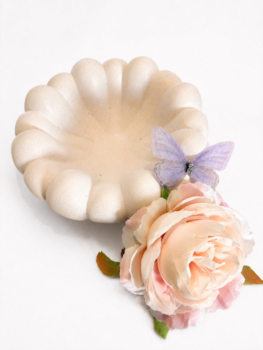 A scalloped cream-colored ceramic dish or shallow bowl, styled with a light pink peony flower and a small purple butterfly on a clean white background.