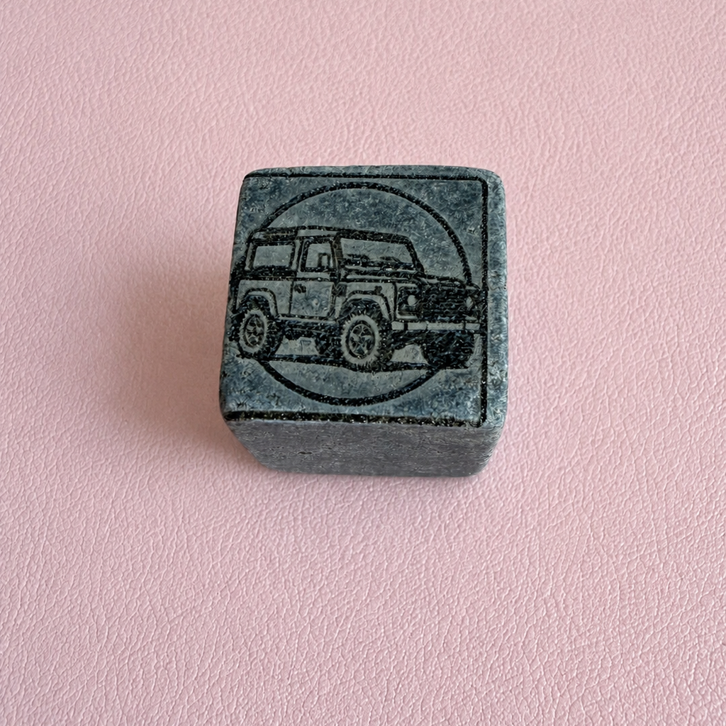 A square dark gray stone or ceramic block featuring an engraved illustration of a vintage off-road vehicle inside a circle, set against a solid light pink background.