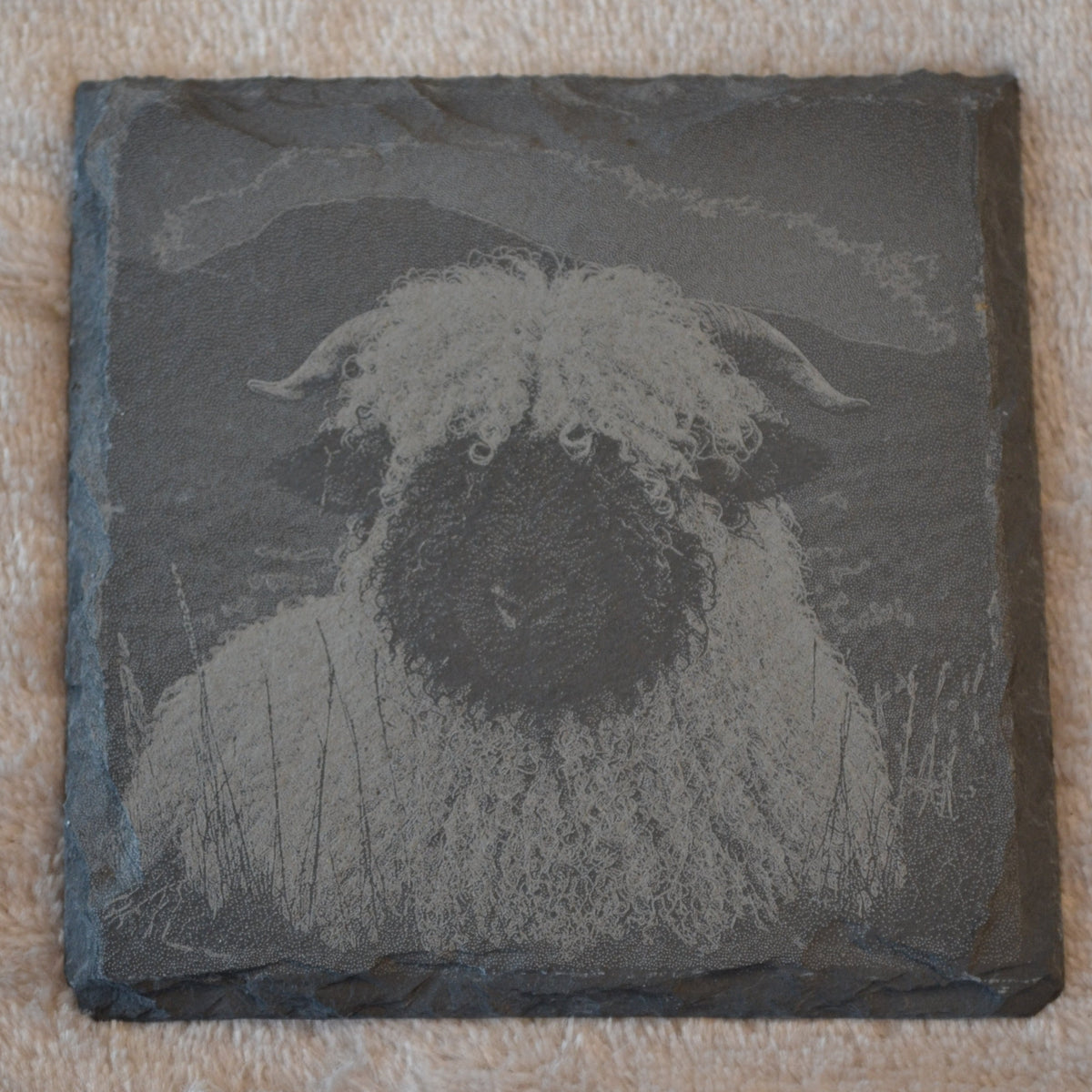 A square dark slate coaster featuring a monochrome engraved or printed portrait of a fluffy dog, placed on a textured beige fabric background.