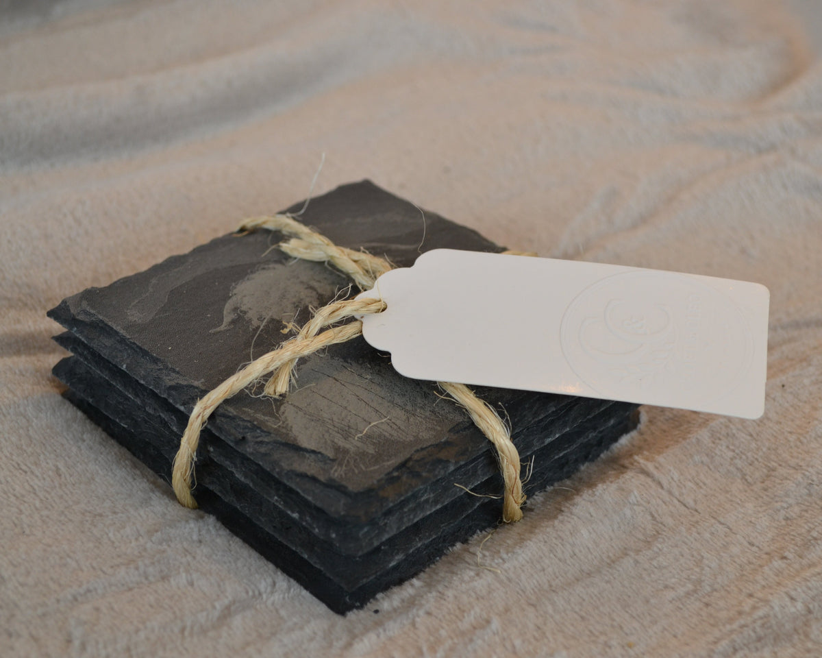 A stack of dark charcoal slate coasters tied with natural jute twine and a blank white paper tag, resting on a textured beige fabric background.