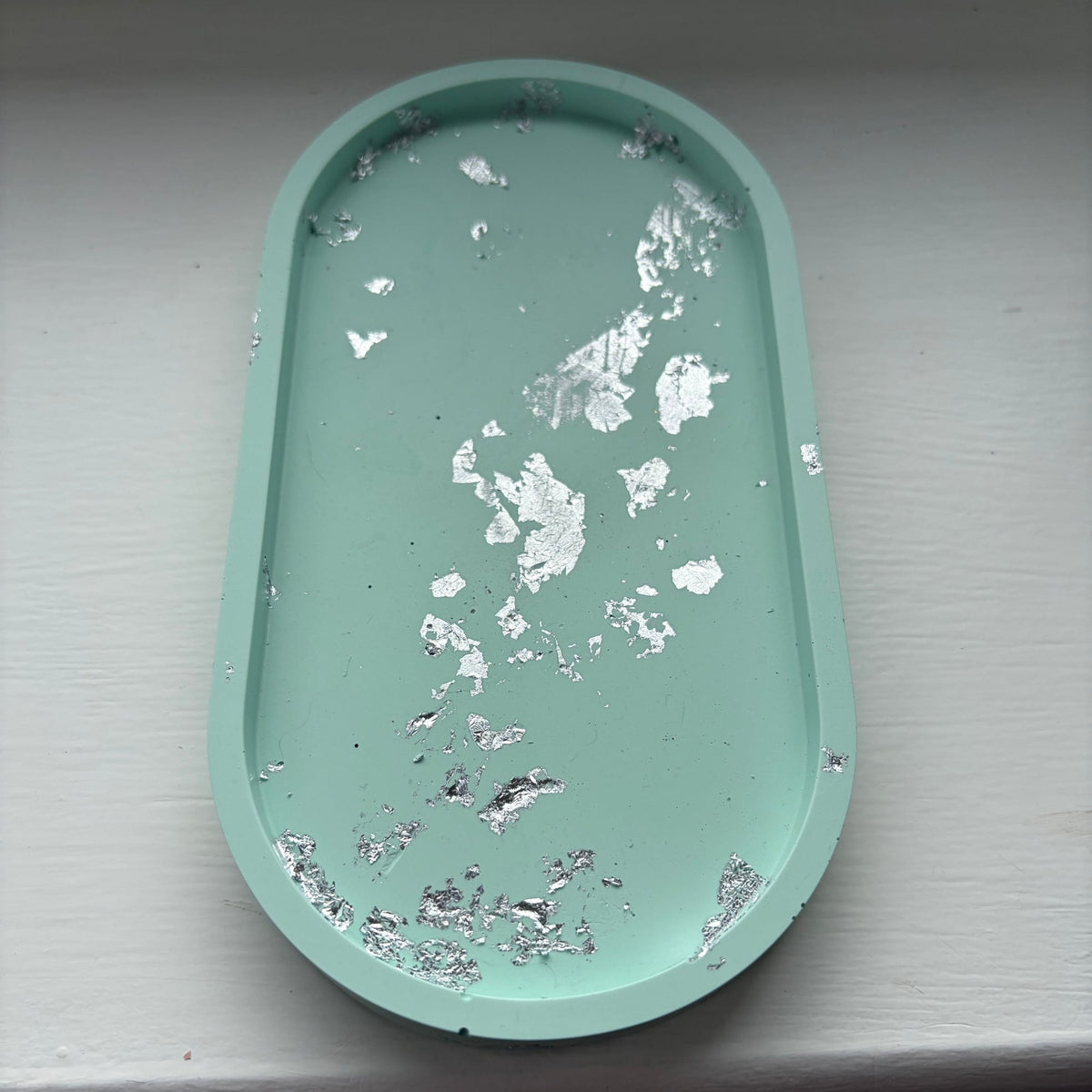 A light green oval jesmonite tray with a distressed silver leaf finish, resting on a white painted wooden surface.