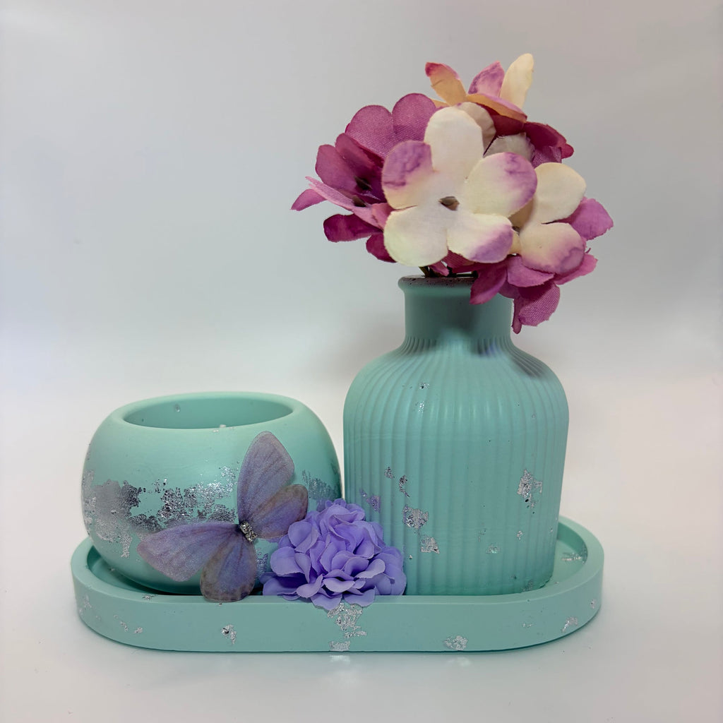 A decorative turquoise set on a white background, featuring a ribbed vase with artificial pink and white flowers, a matching round candle holder with a purple butterfly, and a matching tray.