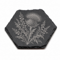 A hexagonal dark gray slate coaster featuring a detailed engraved botanical design of a thistle and surrounding leaves, shown on a plain white background.