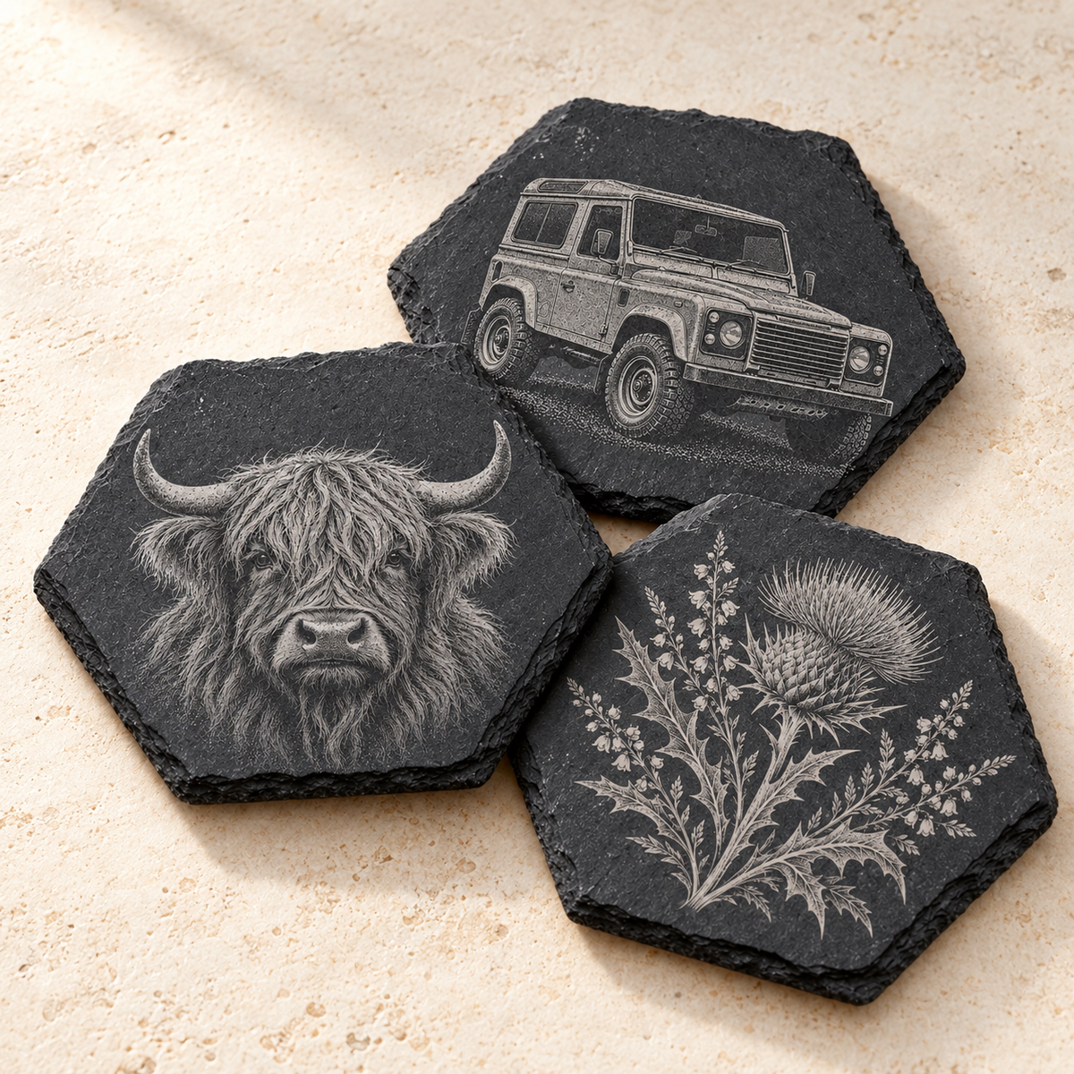 Three hexagonal dark slate coasters with engraved animal and floral designs, arranged on a light beige textured surface. One coaster shows a Highland cow, one shows a vintage Land Rover, and one shows a thistle plant.