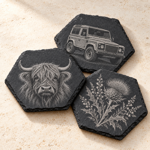 Three hexagonal dark slate coasters with engraved animal and floral designs, arranged on a light beige textured surface. One coaster shows a Highland cow, one shows a vintage Land Rover, and one shows a thistle plant.